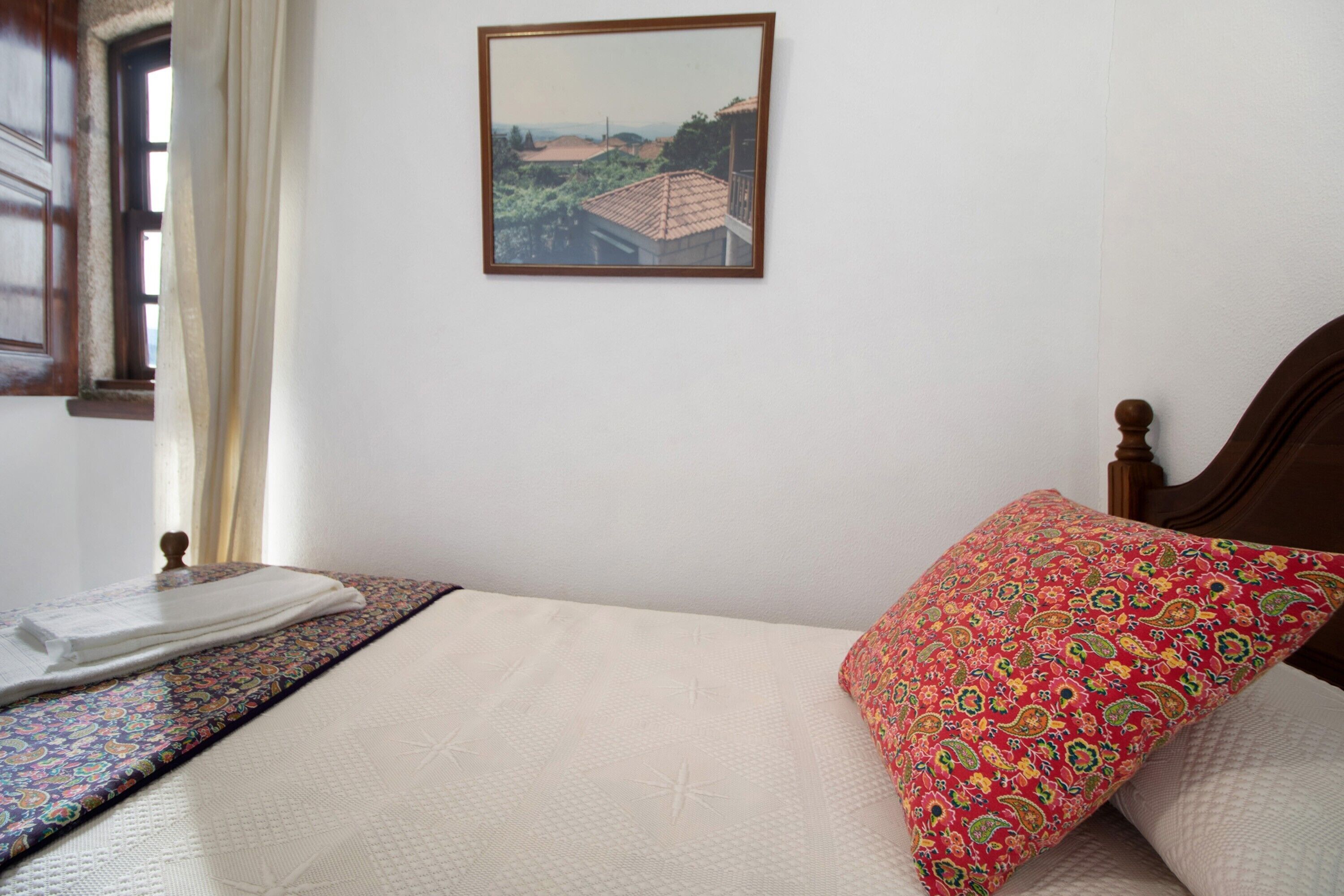 A bed with a patterned bedspread, a decorative pillow, a framed picture on the wall, and a window with curtains.
