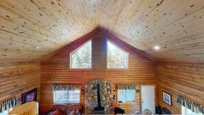 A log cabin interior with a stone fireplace, wooden walls, and two large windows.