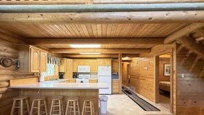 A log cabin interior with a kitchen area featuring wooden cabinets, a central island with stools, and a microwave.