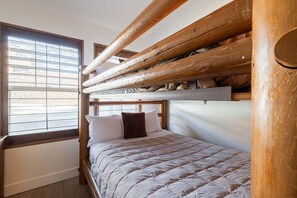 Un lit superposé avec une tête de lit en bois, une fenêtre avec des stores et une couvre-lit matelassé.