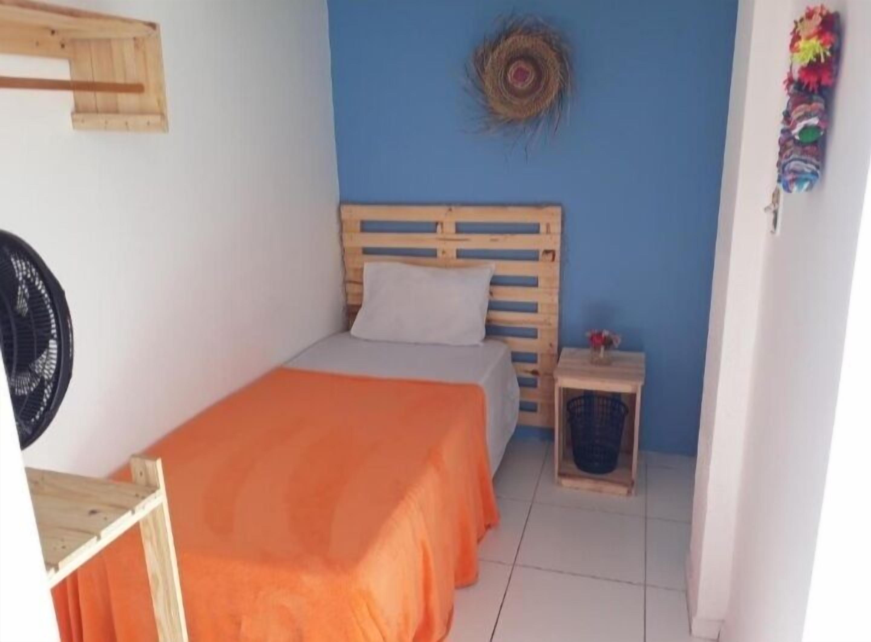 A small, simple room with a wooden bed, a small table, and a fan.