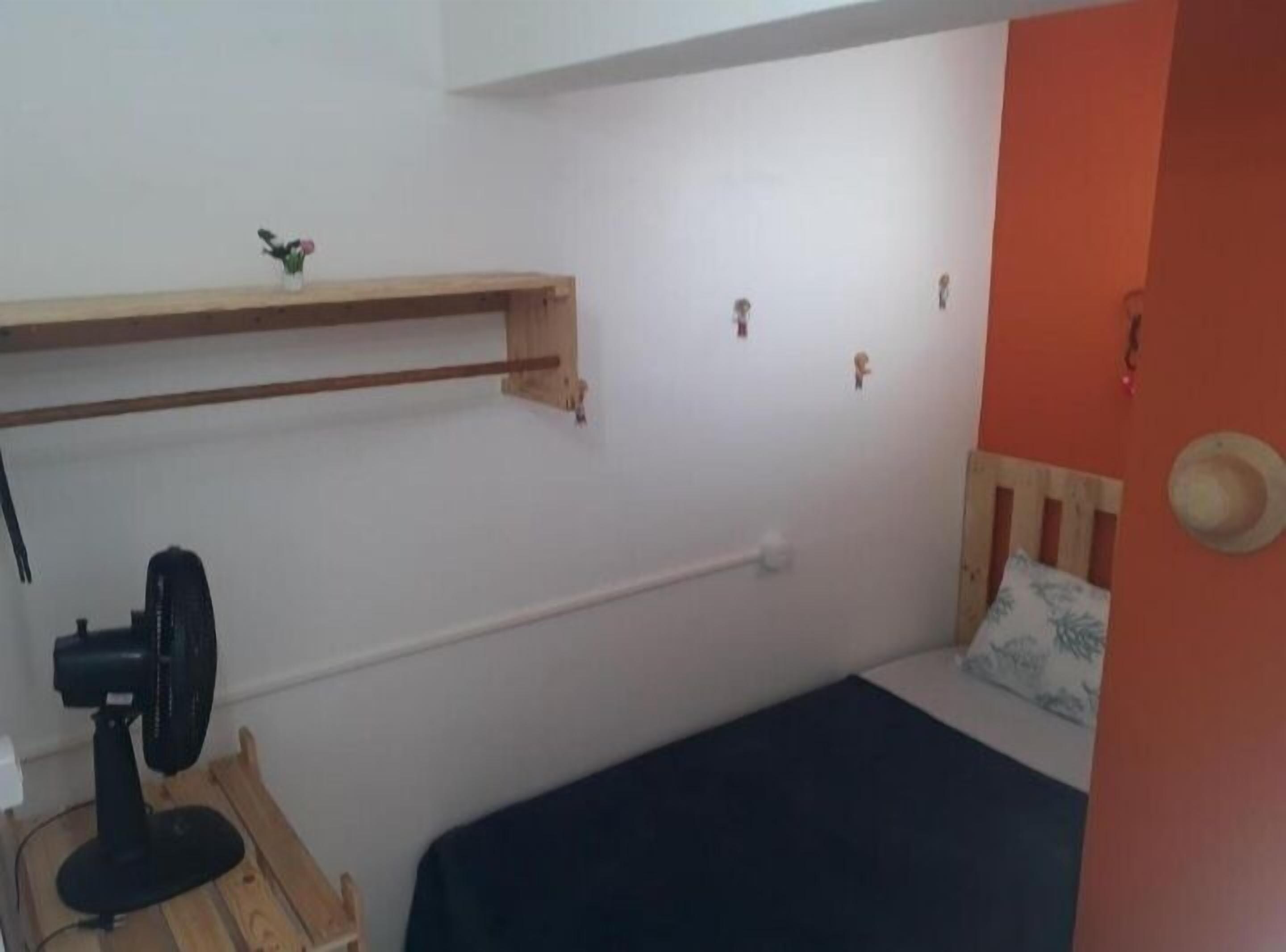 Pequeña habitación funcional con una cama individual, un estante de madera, un escritorio con silla y un ventilador de pared.