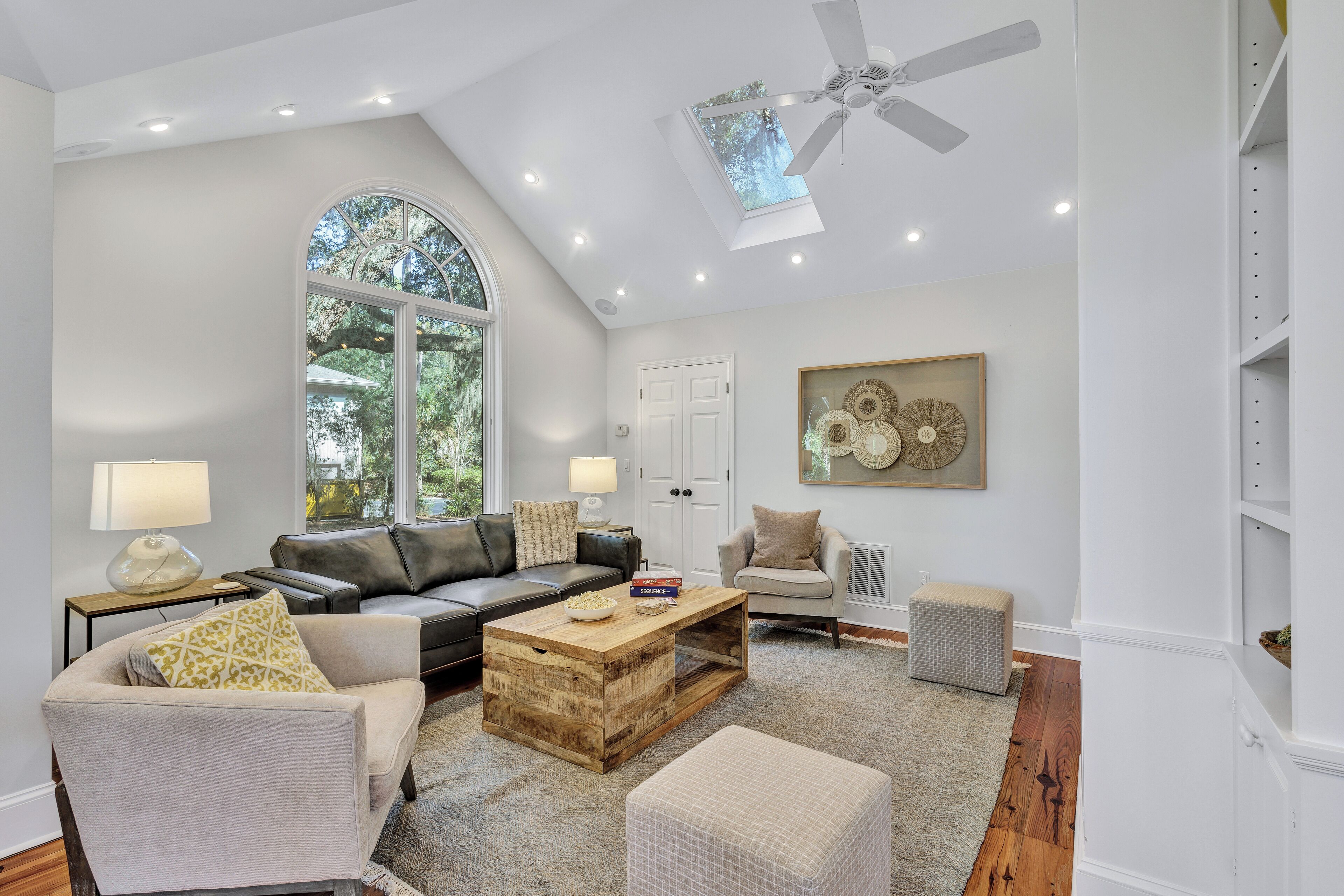 A spacious living room with a high ceiling, a large window, a ceiling fan, and a wooden coffee table.