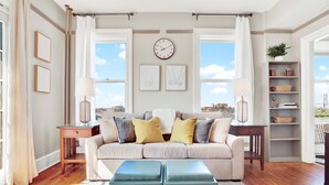 Un salon avec un canapé beige, deux tables d’appoint, une horloge, des œuvres d’art encadrées et une vue dégagée grâce à de grandes fenêtres.