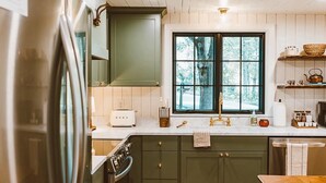 Una cocina moderna con pisos de madera, gabinetes verdes, un refrigerador de acero inoxidable y un ventanal con vista a árboles.