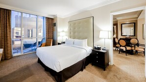 Habitació d'hotel amb un llit gran, una taula de nit amb làmpada, una cadira, una taula de menjador amb caires i una finestra gran amb cortines.