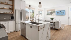 Una cocina moderna con armarios blancos, una isla de mármol, electrodomésticos de acero inoxidable y un comedor con sofá y planta.