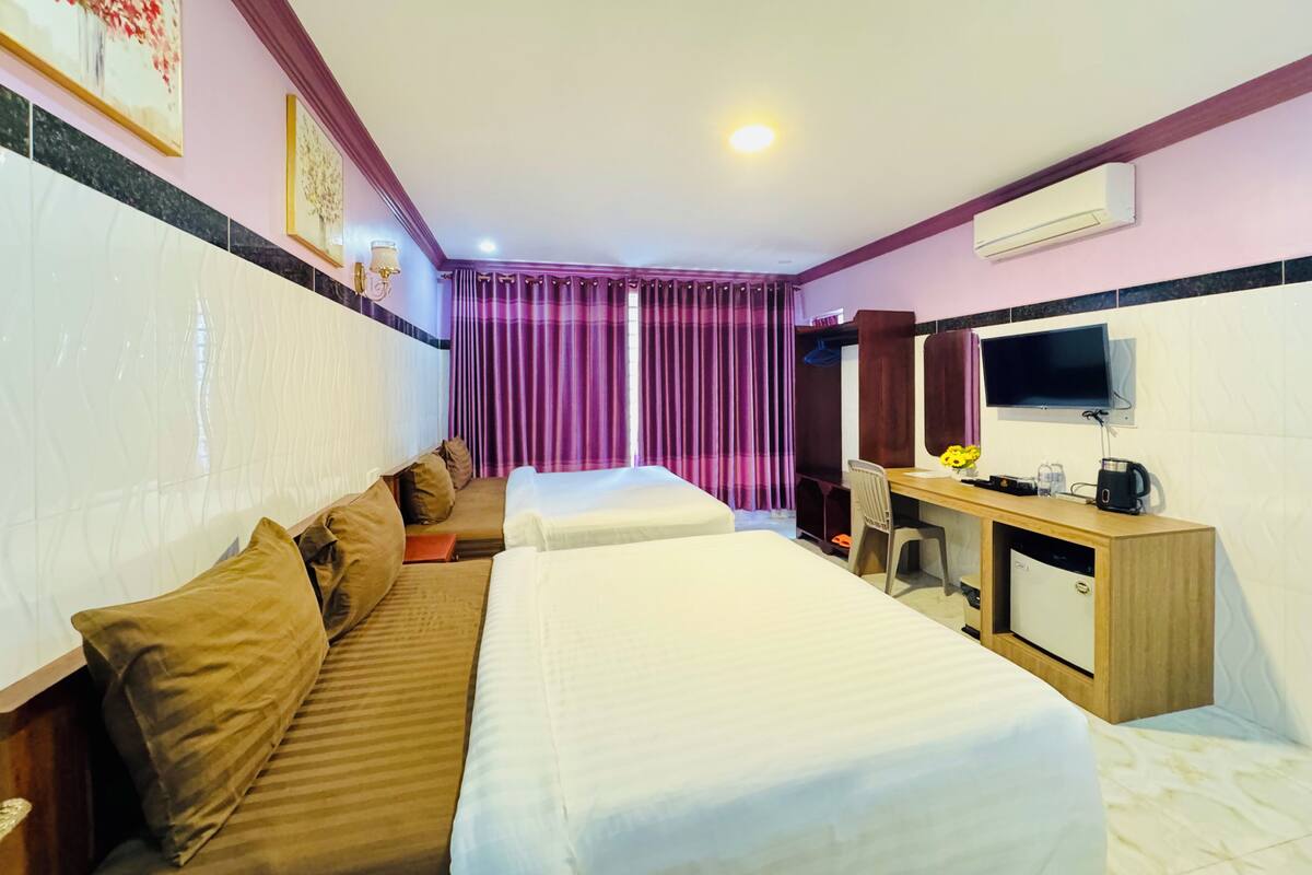 Ein Hotelzimmer mit zwei Betten, einem Sofa, einem Schreibtisch mit Stuhl, einem Fernseher und einer Klimaanlage.