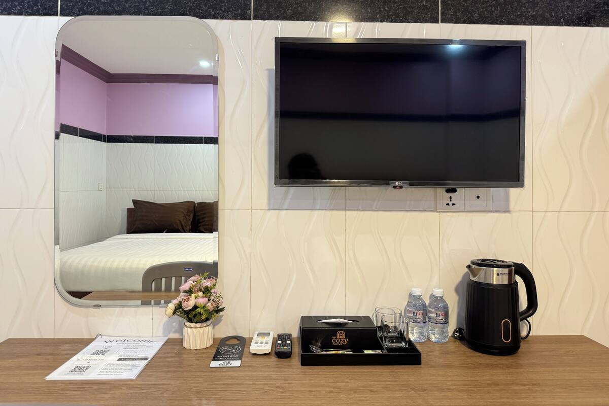 Ein Hotelzimmer mit einem Bett, einem an der Wand montierten Fernseher, einem Spiegel, einem Tisch mit Wasserkocher, Gläsern, einer Wasserflasche, einer Fernbedienung und einer Willkommensbroschüre.