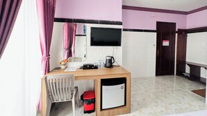 A hotel room with a wooden desk, a television mounted on the wall, a small refrigerator, a trash bin, and a door with a sign. - The Cozy Corner-Budget Hotel (Kampot)