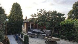 En träaltan med utemöbler, omgiven av grönska och en byggnad i bakgrunden.
