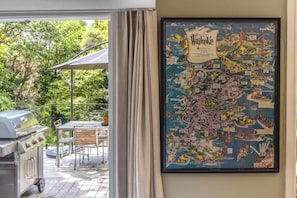 A framed map of Waikiki on a wall, a grill, and a patio table with chairs. - Hillside Hideaway – Magical Cottage Escape (Waiheke Island)