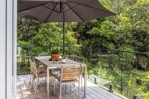 Une terrasse en bois avec une table et des chaises, une plante en pot et une bouteille de vin, le tout entouré d’une végétation luxuriante.