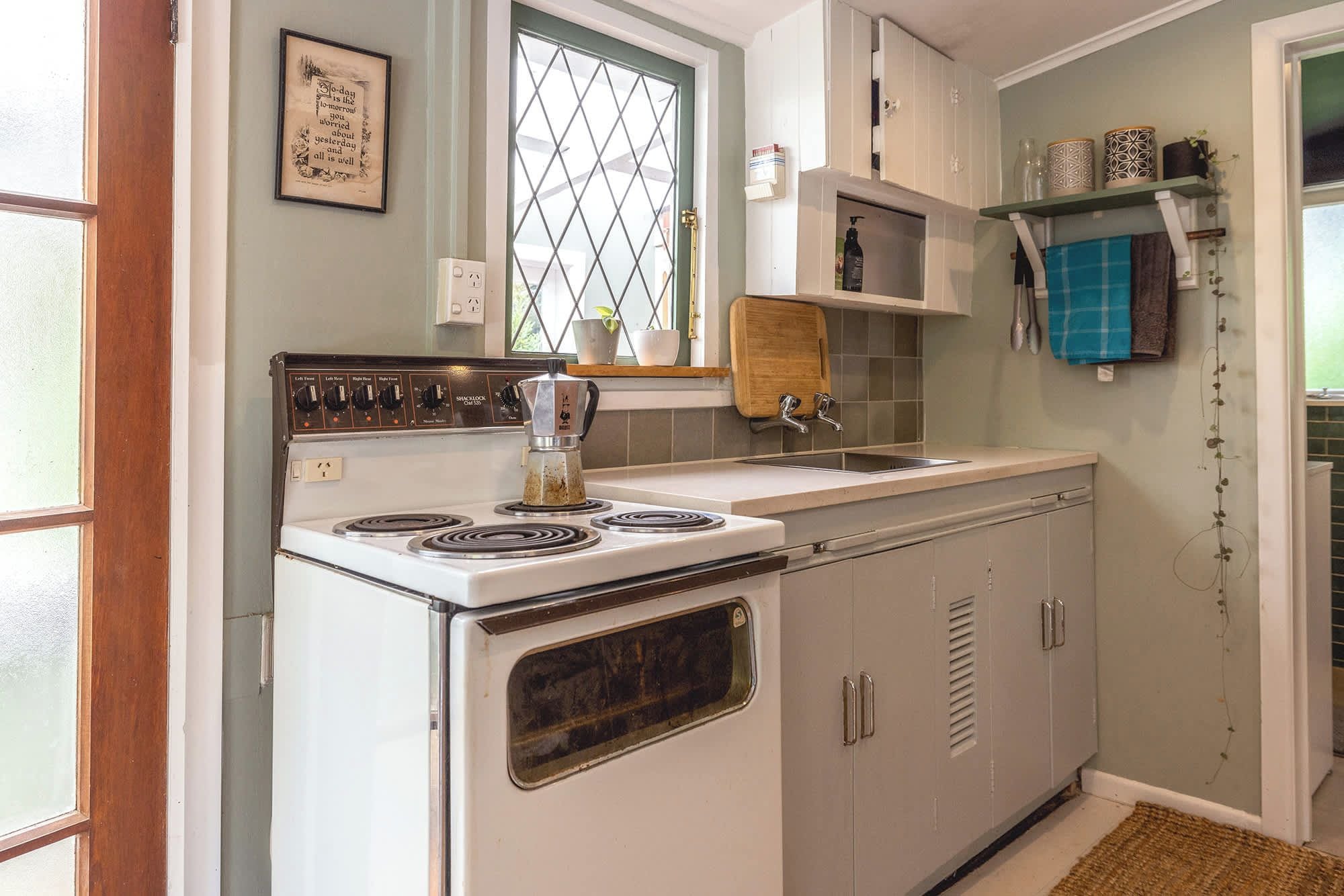 A kitchen with a white stove, a sink, and cabinets. There is a window with a diamond pattern, a wooden cutting board, and a shelf with kitchen items.