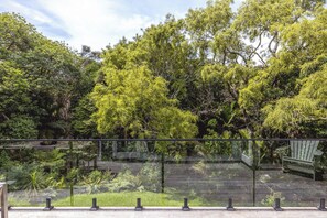 Un balcon doté d’une balustrade en verre, donnant sur un jardin luxuriant où poussent différentes espèces d’arbres et d’arbustes.