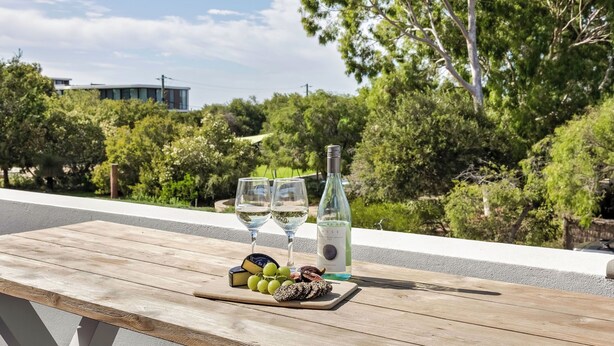 Une table en bois, une bouteille de vin blanc, deux verres, ainsi qu’un assortiment de raisins et de fromages, sont disposés sur un balcon donnant sur un paysage verdoyant et luxuriant, avec des arbres et un bâtiment en arrière-plan.