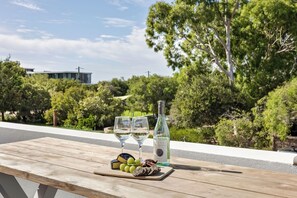 A wooden table with a bottle of white wine, two glasses, and a selection of grapes and cheese on a balcony overlooking a lush green landscape with trees and a building in the background. - Salty Solstice - Beachside Dunsborough Town  (Dunsborough)