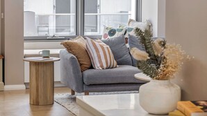 Un salon moderne avec un canapé gris, une petite table d’appoint ronde en bois et un vase blanc contenant des fleurs séchées.