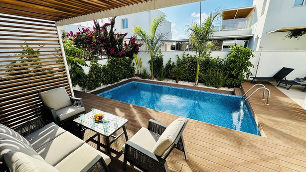 Ein Poolbereich mit Holzterrasse, Gartenmöbeln und Blick auf ein modernes Gebäude im Hintergrund.