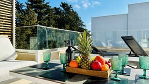 Ein Tisch mit Obstkorb, grünen Gläsern und einer Flasche auf einem Balkon mit Blick auf Bäume und ein Gebäude.