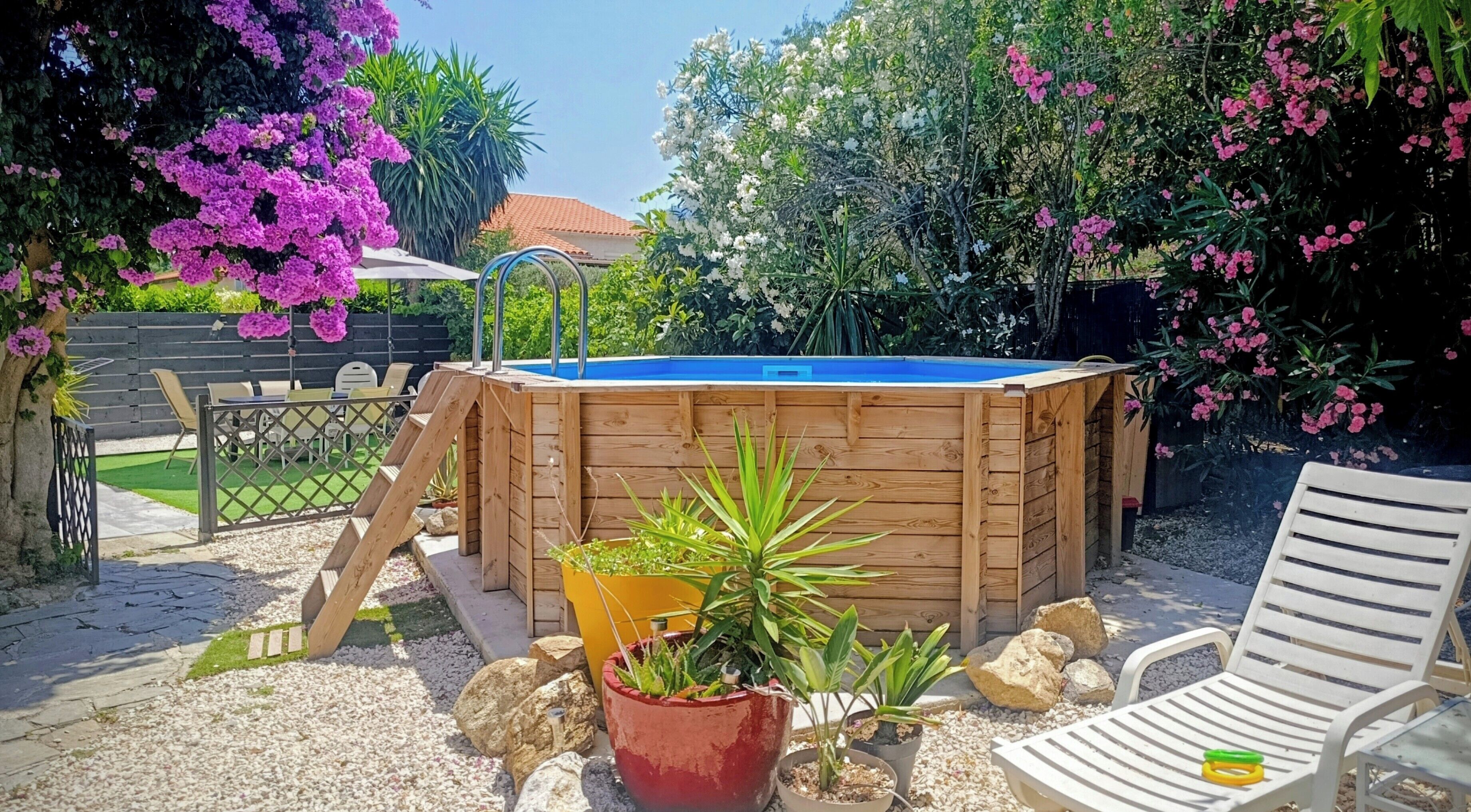 A wooden hot tub with a blue cover, surrounded by potted plants, a white chair, and a patio area with a fence and greenery.