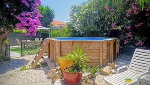 A wooden hot tub with a blue cover, surrounded by potted plants, a white chair, and a patio area with a fence and greenery.