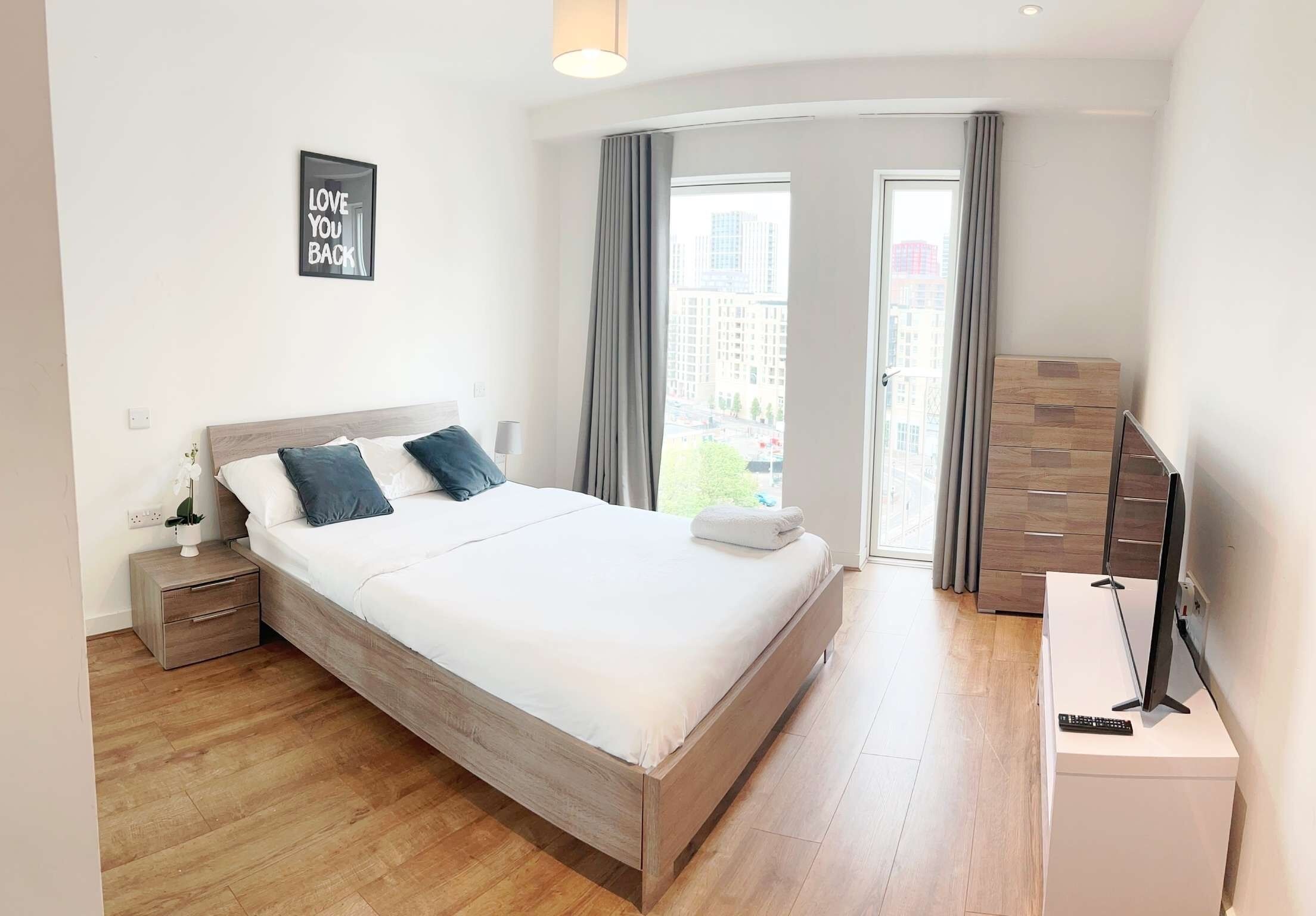 A modern bedroom with a large bed, a bedside table, a TV on a stand, and a window with curtains.