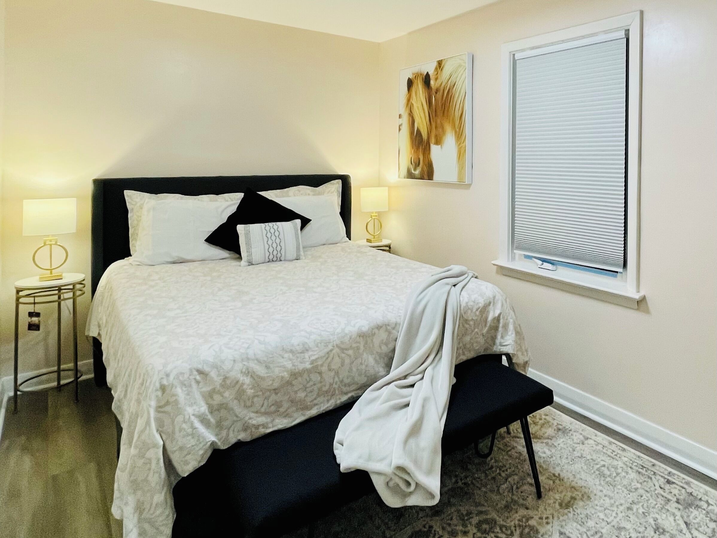 A neatly made bed with a black headboard, two bedside lamps, and a window with blinds.