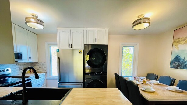 A modern kitchen with white cabinets, a stainless steel refrigerator, a black stove, and a dining area with a wooden table and chairs. - Entire house 3BR, Pets, kitchen, Parking, Laundry, Large Fenced backyard, WiFi (Kitchener)