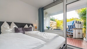 A modern hotel room with a large bed, a nightstand, a window with curtains, and a view of the outdoor seating area. - This one love - Cozy vacation apartment for up to 3 people in Alt (Sylt)