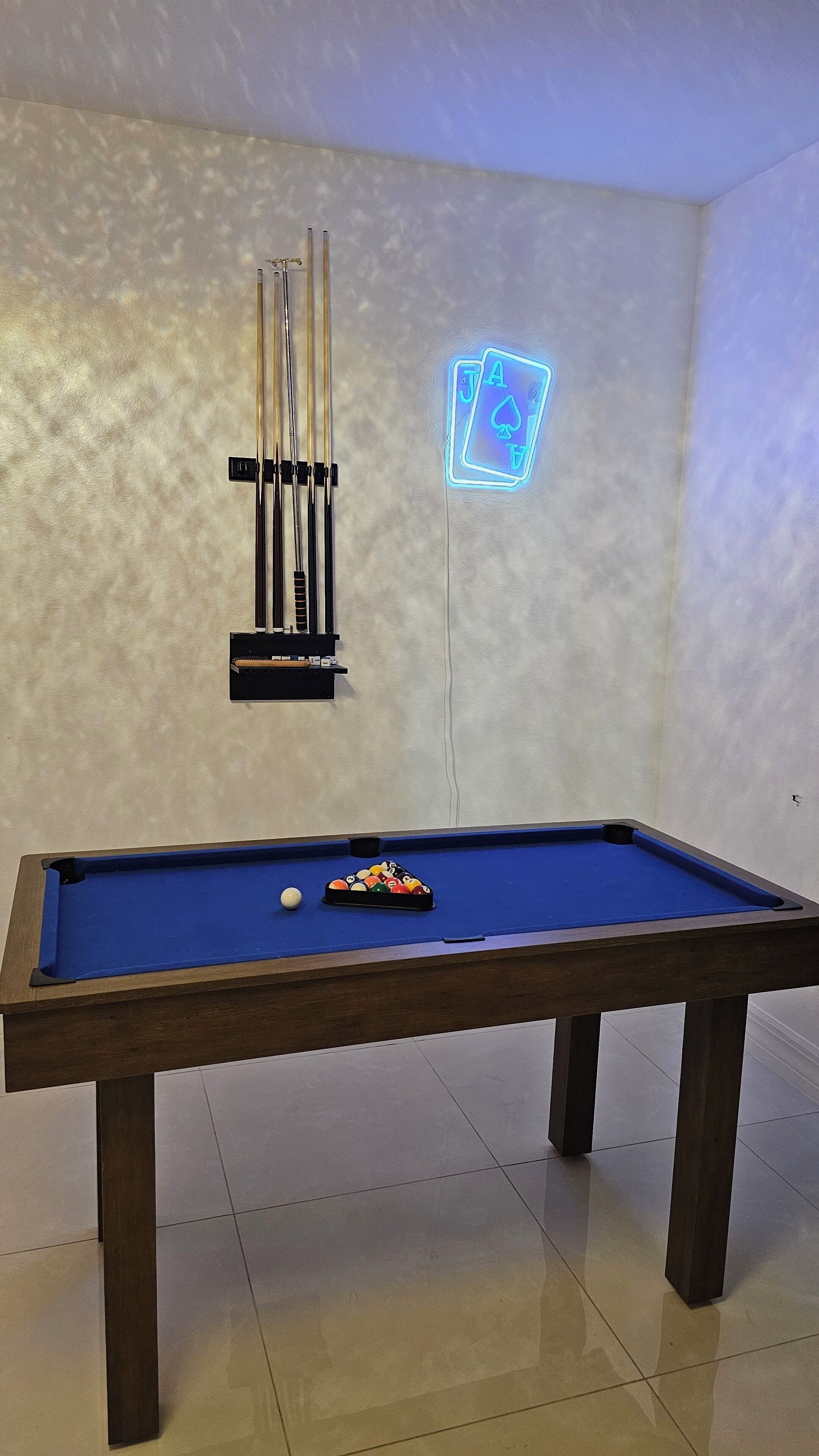 A pool table with a blue felt surface, a set of pool cues, and a neon sign on the wall.