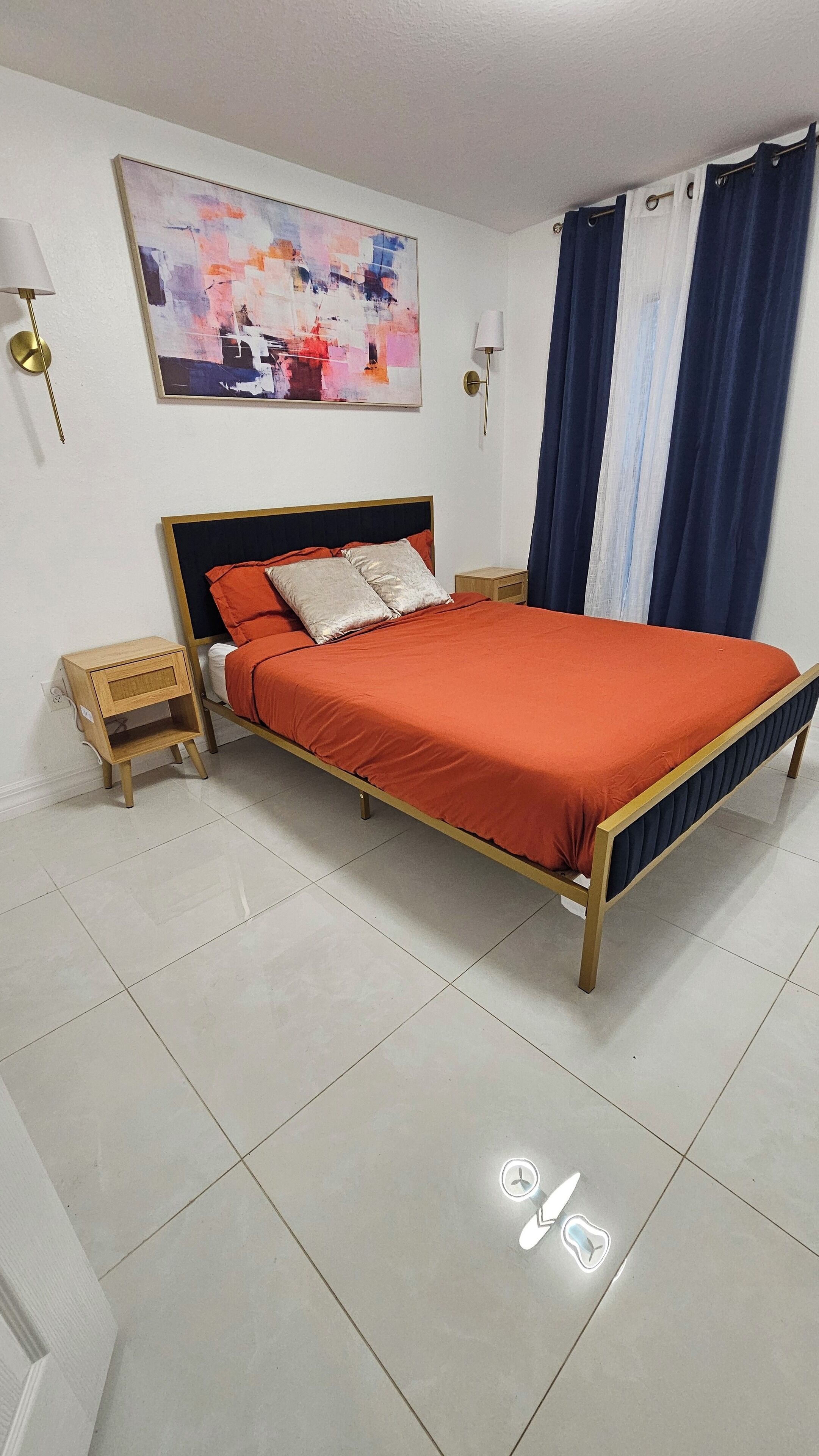 A bedroom with a bed, nightstands, a wall-mounted lamp, a painting, and a window with curtains.