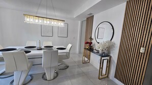 A modern dining room with a white table, black plates, and white chairs. There is a chandelier above the table and a decorative mirror on the wall.