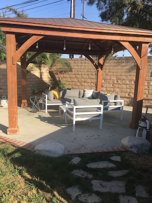 A covered outdoor seating area with a brick wall backdrop, a wooden pergola, and a white sofa set. - 3-bedroom house in East Whittier with AC/heating for a relaxing stay any season! (Whittier)