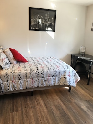 Une chambre à coucher avec un lit, un tableau représentant un paysage urbain encadré, une petite table avec un panier, et un plancher en bois.
