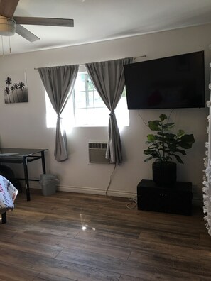A room with a flat-screen TV mounted on the wall, a ceiling fan, a window with curtains, a potted plant, a table, and a trash bin. - 3-bedroom house in East Whittier with AC/heating for a relaxing stay any season! (Whittier)