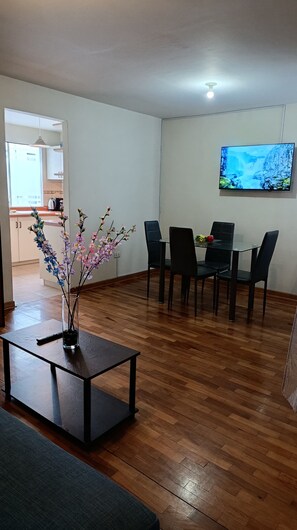 Ein Wohnzimmer mit Essbereich, Holzboden, einem Glastisch und einem an der Wand montierten Fernseher.
