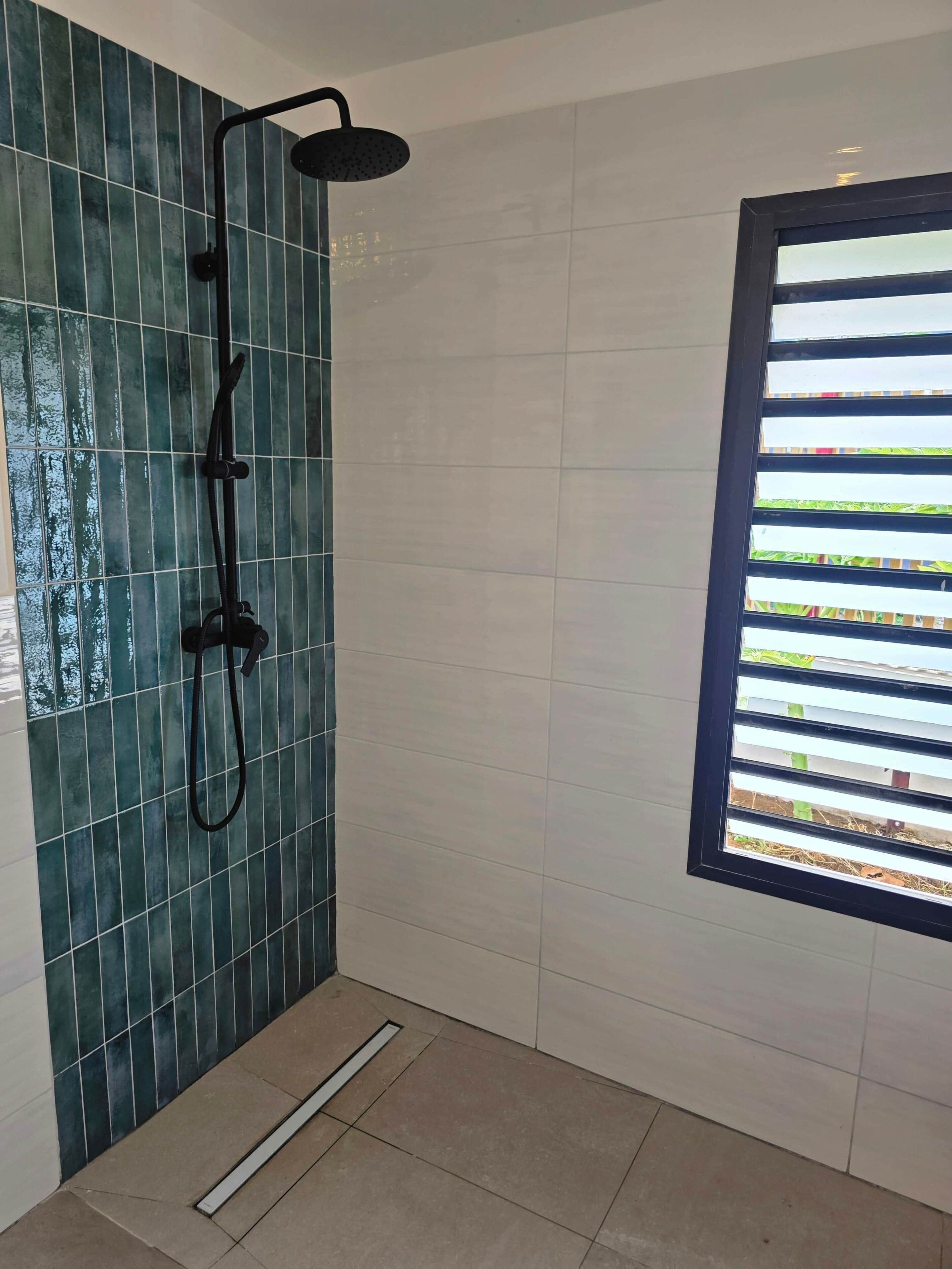 Une douche équipée d’un pommeau de douche à effet pluie et d’un pommeau de douche à main, une fenêtre avec des stores horizontaux et un sol carrelé.