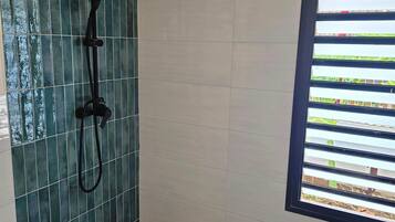 Une douche équipée d’un pommeau de douche à effet pluie et d’un pommeau de douche à main, une fenêtre avec des stores horizontaux et un sol carrelé.