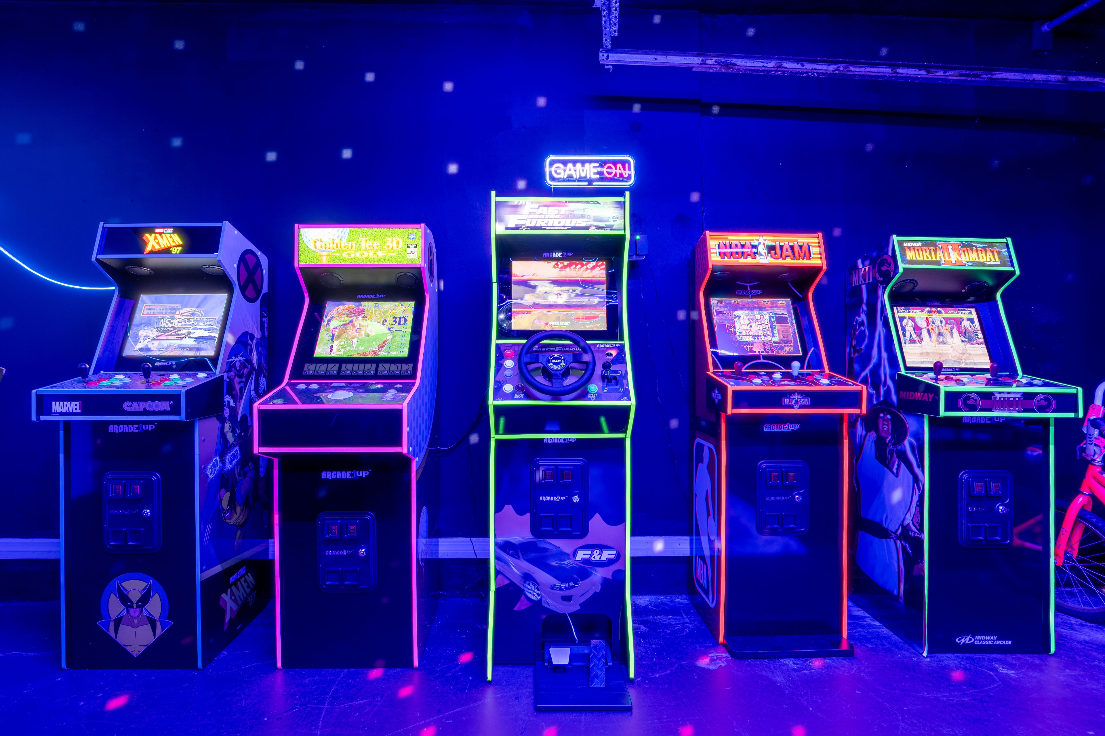 Four arcade machines with neon signs, including games like 'X-Men' and 'Mortal Kombat'.