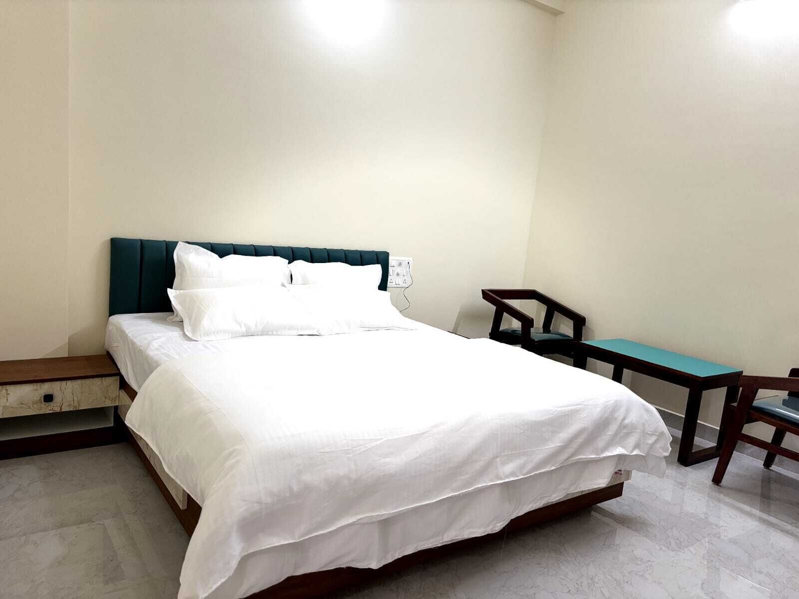 Una cama bien tendida con sábanas blancas y un cabecero color turquesa, una mesita de noche de madera, una pequeña mesa con una silla y un enchufe.