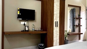 A bedroom with a bed, a TV mounted on a wooden cabinet, a small shelf with toiletries, and a potted plant. - Evara Enclave Residency (Sullia)