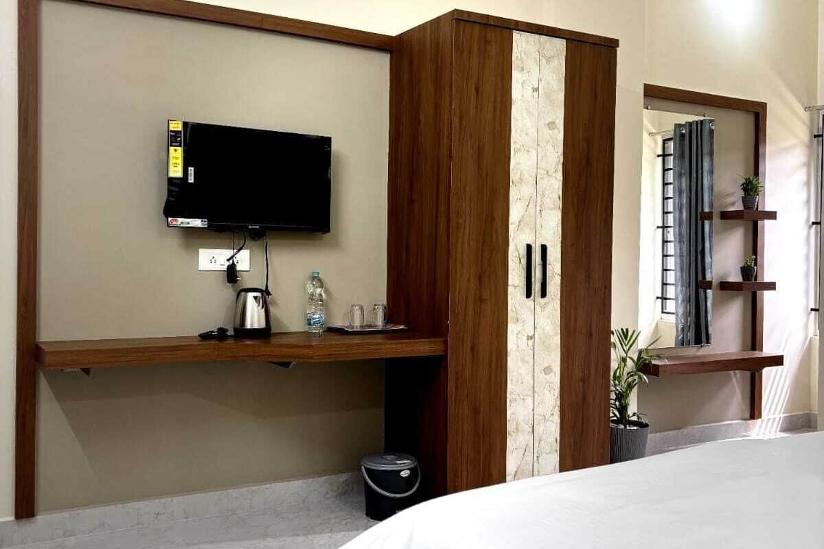 A bedroom with a bed, a TV mounted on a wooden cabinet, a small shelf with toiletries, and a potted plant.