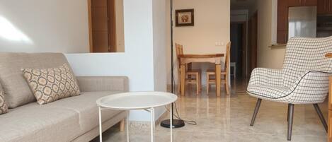 A living room with a sofa, a small round table, a dining table with chairs, and a kitchen area with cabinets.