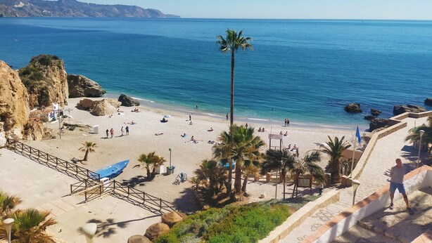 Ein Strand mit klarem, blauem Wasser, Palmen und einer felsigen Klippe.