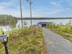 A gravel path leading to a wooden dock over a calm lake, surrounded by coniferous trees and a clear sky.