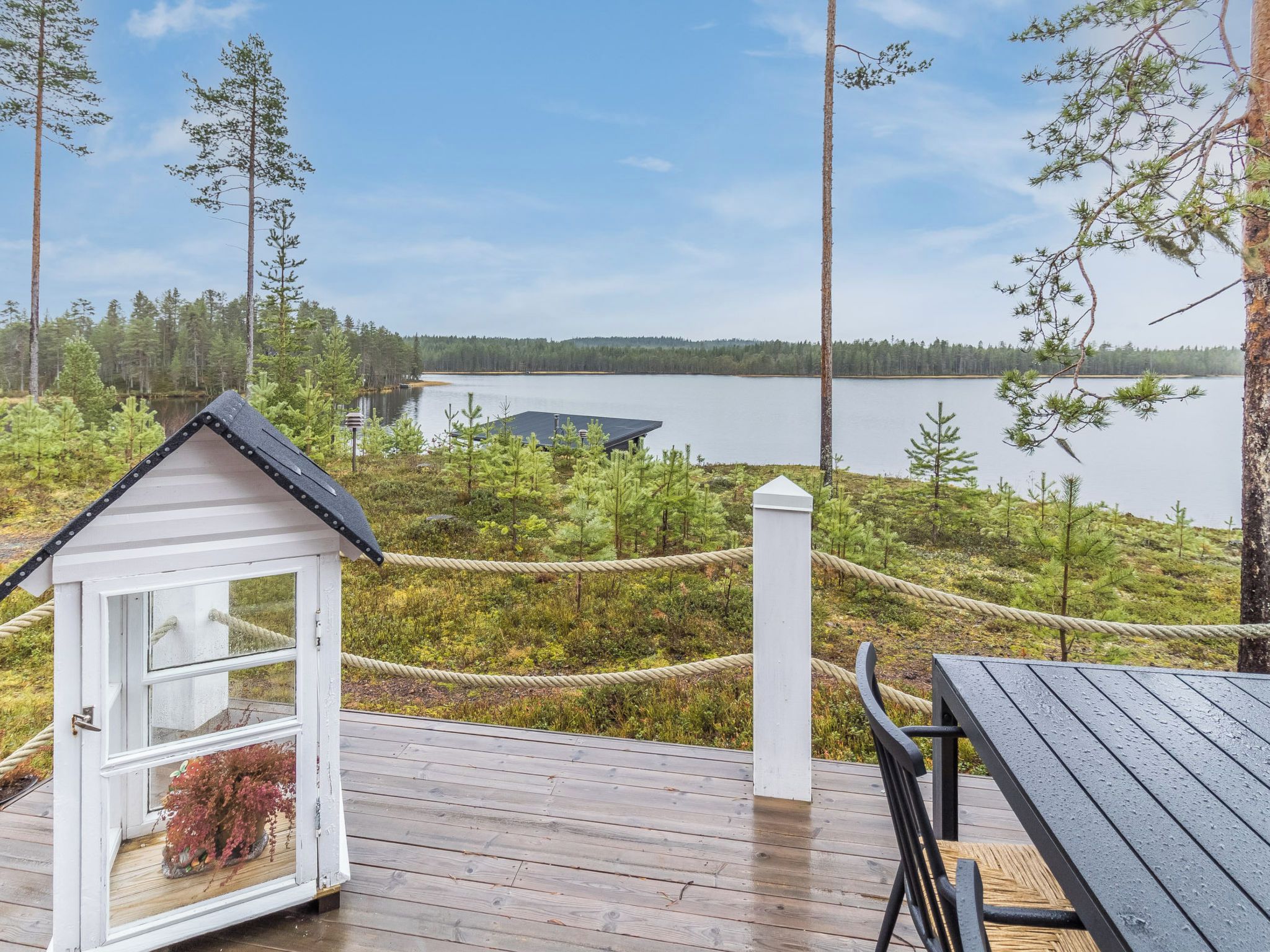 A wooden deck with a white cabin-like structure and a table, overlooking a calm lake surrounded by trees.