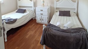 Quarto com duas camas de solteiro, mesa de cabeceira branca, luminária pendente azul-esverdeada e piso de madeira.