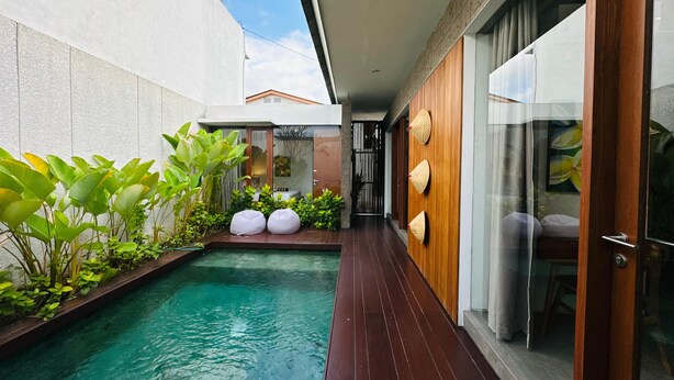 A modern outdoor pool area with a wooden deck, surrounded by lush greenery and a glass door leading to a room.