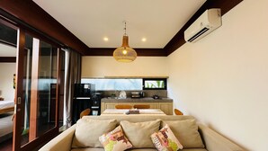 A modern living room with a beige sofa, two decorative pillows, a hanging light fixture, and a kitchen in the background.